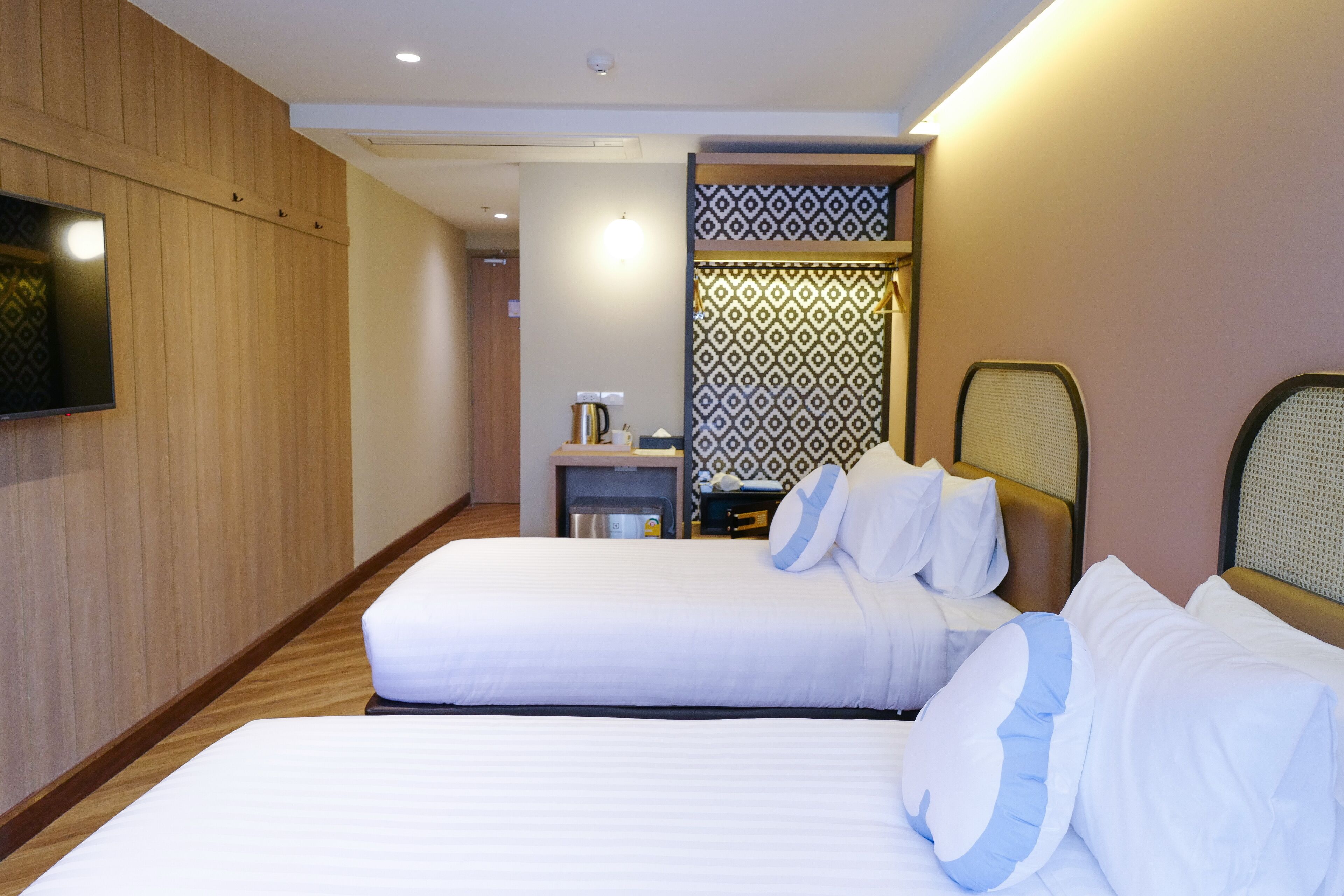 A hotel room with two beds, a TV mounted on the wall, a small table with a kettle, and a patterned wall divider.