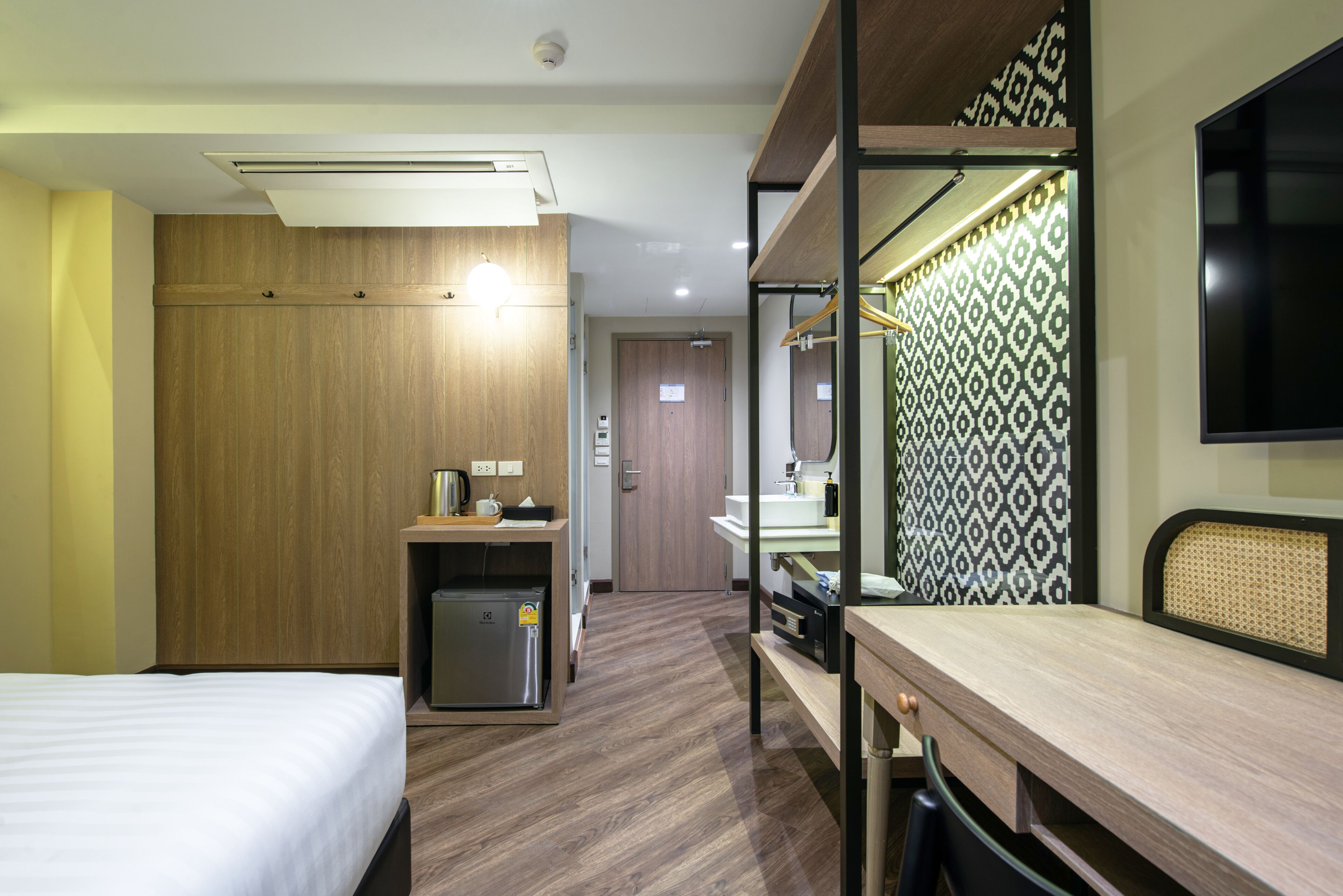 A hotel room with a bed, a desk, a wardrobe, a small fridge, a mirror, and a patterned wall.