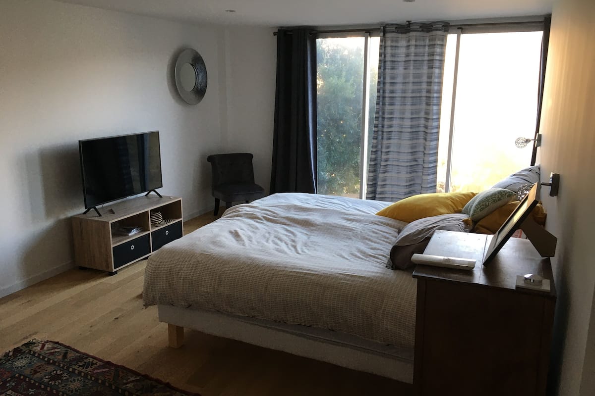 Ein Schlafzimmer mit einem Bett, einem Fernseher auf einem Holzboden, einem Sessel und einem Fenster mit Vorhängen.