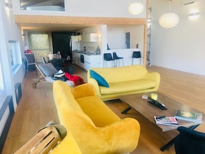 Ein geräumiger Wohnbereich mit einem gelben Sofa, einem Holztisch und einem Esstisch. Im Hintergrund ist eine Küche mit modernen Geräten und Barhocker zu sehen.