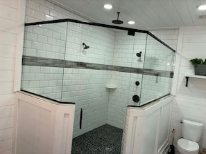 Un baño en esquina con paredes de azulejos blancos, un box de ducha de cristal, piso de baldosas negras y un inodoro blanco.