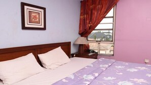 Quarto com cama, travesseiros, mesa de cabeceira, luminária, quadro emoldurado e janela com cortinas.