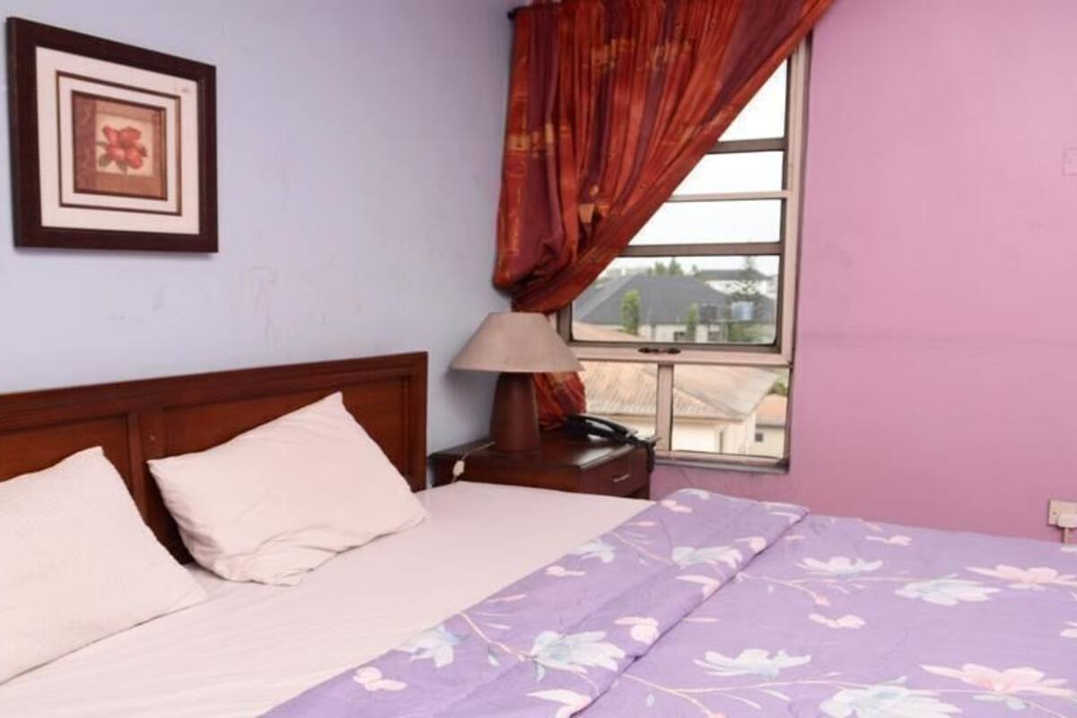Une chambre à coucher avec un lit, une table de chevet, une lampe, un tableau encadré au mur et une fenêtre avec des rideaux.