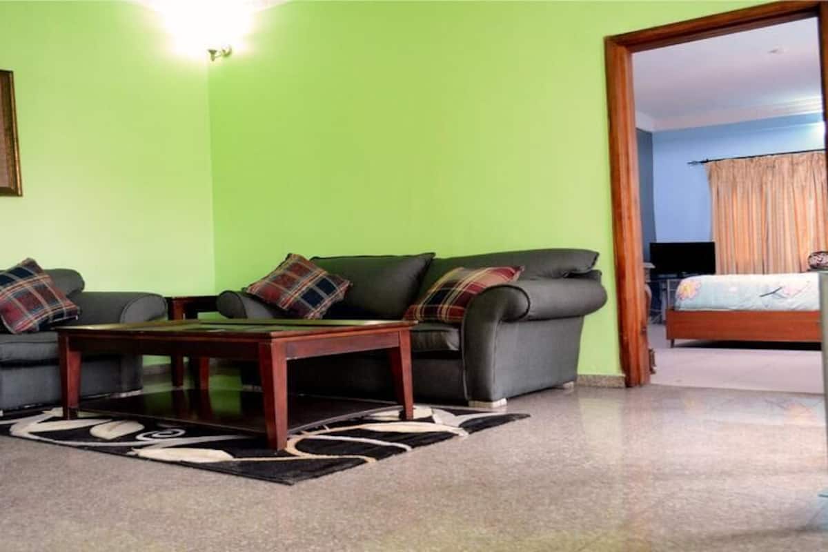 Un salon avec un mur vert, un ensemble de canapés, une table basse et une vue sur une chambre à coucher avec un lit et des rideaux.
