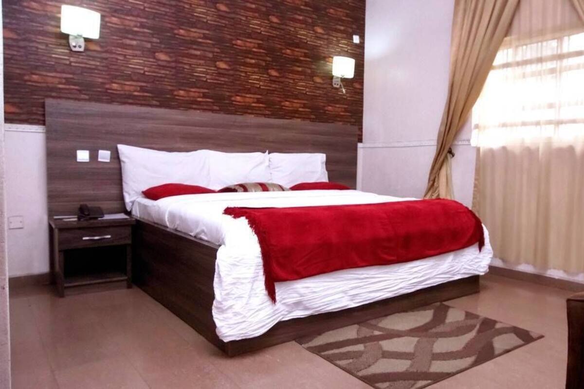A bedroom with a wooden headboard, a large bed with white and red bedding, a nightstand, and a window with curtains.
