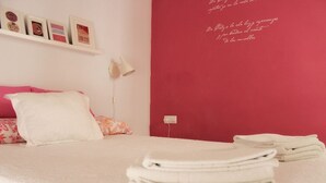 Cama bem arrumada com lençóis brancos, um travesseiro rosa e uma parede vermelha com texto decorativo.