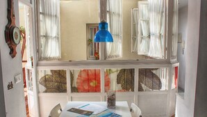 Uma área de refeições com uma mesa posta para dois, uma luminária pendente azul e vista para um jardim através de uma janela com cortinas brancas.