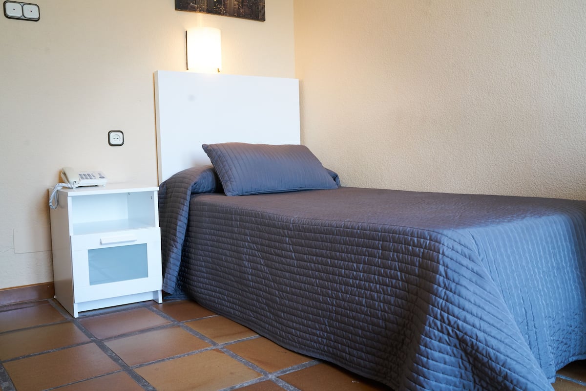 A single bed with a grey quilt, a white bedside table with a phone, and a wall socket.