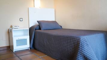 Una cama individual con un edredón gris, una mesita de noche blanca con un teléfono y un enchufe de pared.