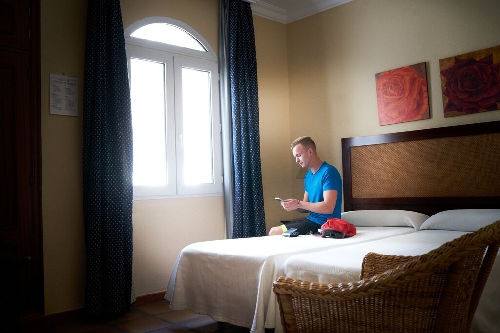 Een hotelkamer met een bed, een rieten stoel, een raam met gordijnen en twee schilderijen aan de muur.