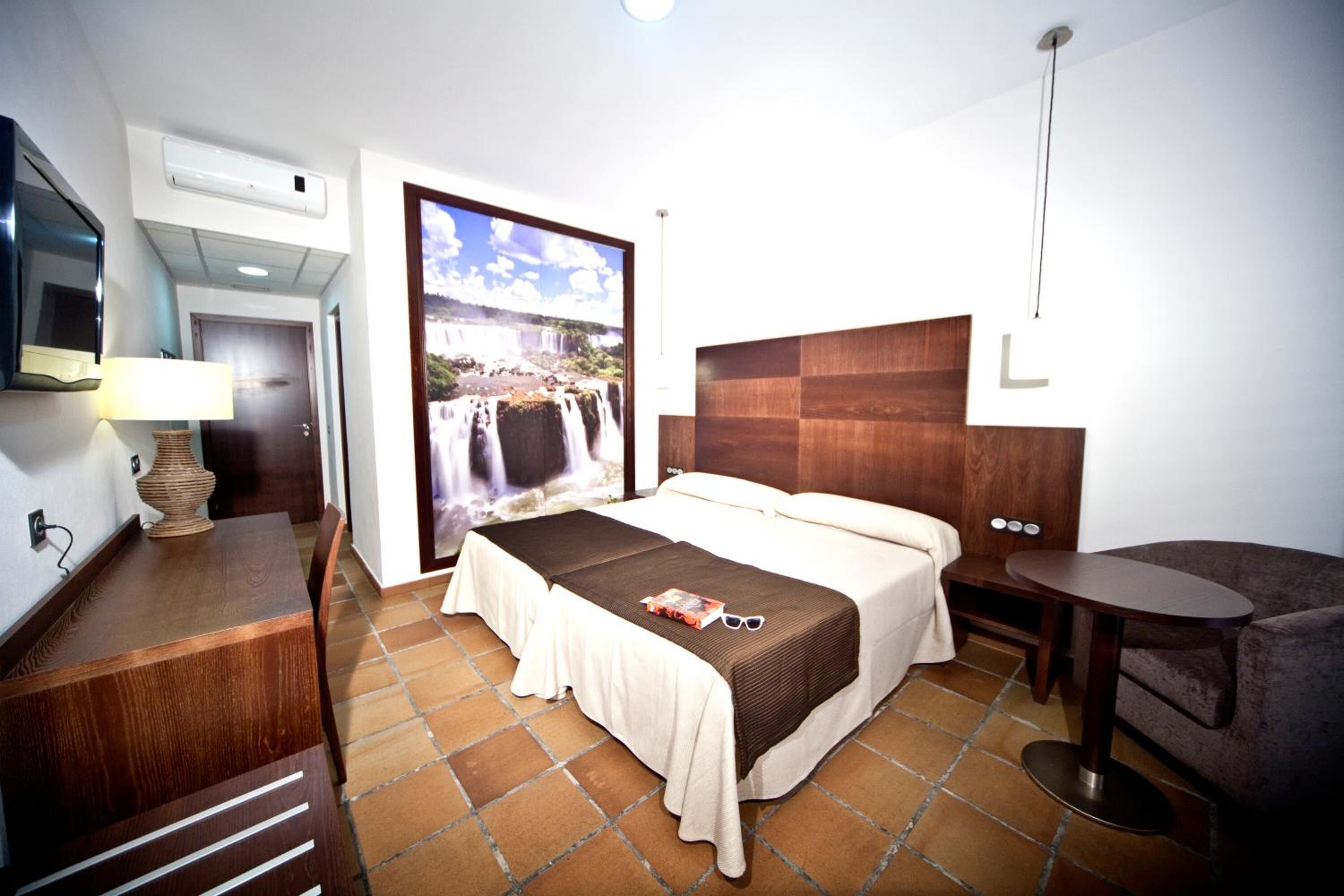 Habitació d'hotel amb llit, escriptori, cadira, televisió i una finestra gran que dona a una cascada.