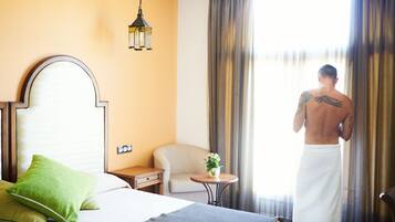 A hotel room with a bed, a chair, a small table with a plant, and a window with curtains.