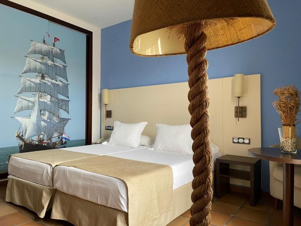 Camera d'albergo con un letto grande, una testiera con una lampada, un quadro raffigurante una nave sulla parete e un tavolino con un vaso.