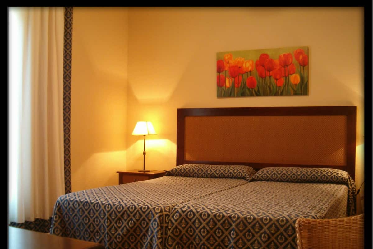A hotel room with a patterned bedspread, a painting of red flowers on the wall, and a bedside lamp.