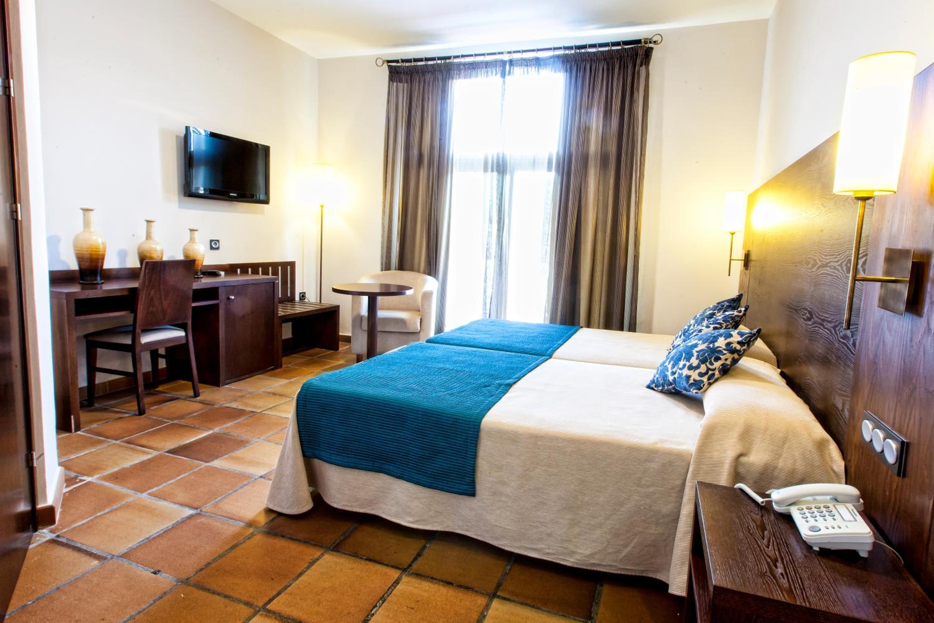 Habitación de hotel con cama, mesita de noche, lámpara, escritorio, silla, televisor y ventana con cortinas.