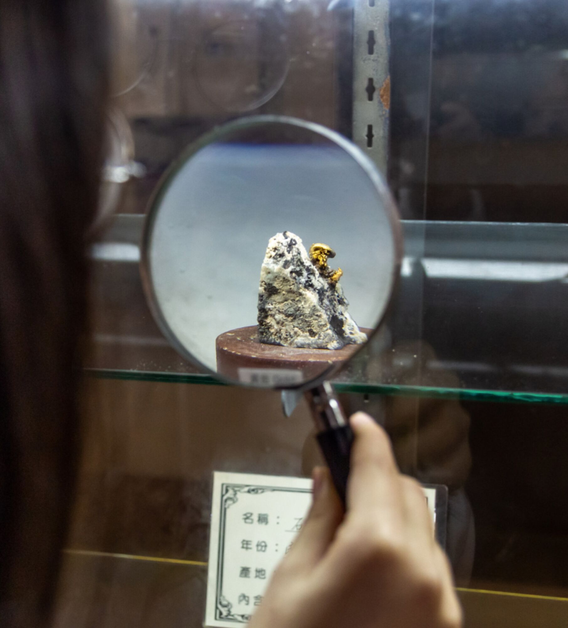 A hand holding a magnifying glass focusing on a rock specimen in a display case.