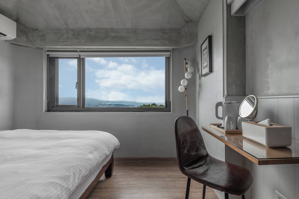 Un dormitorio con cama, escritorio y silla, ventana con vista y espejo instalado en la pared.