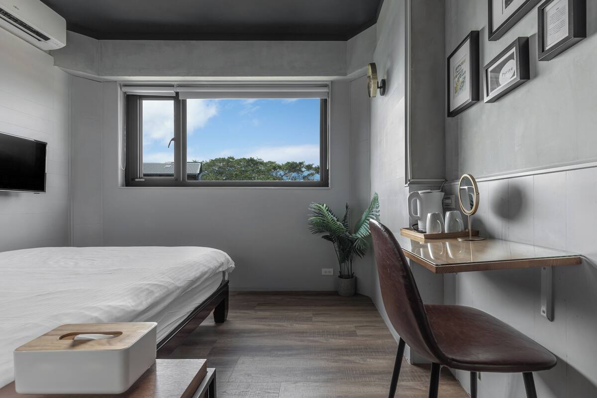 Un dormitorio con cama, escritorio y silla, una planta en maceta y ventana con vistas a árboles.