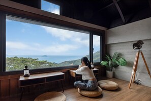 A room with a large window, wooden furniture, a small table with a lantern, a potted plant, and an easel. - Jiufen The Ocean Theory (New Taipei City)