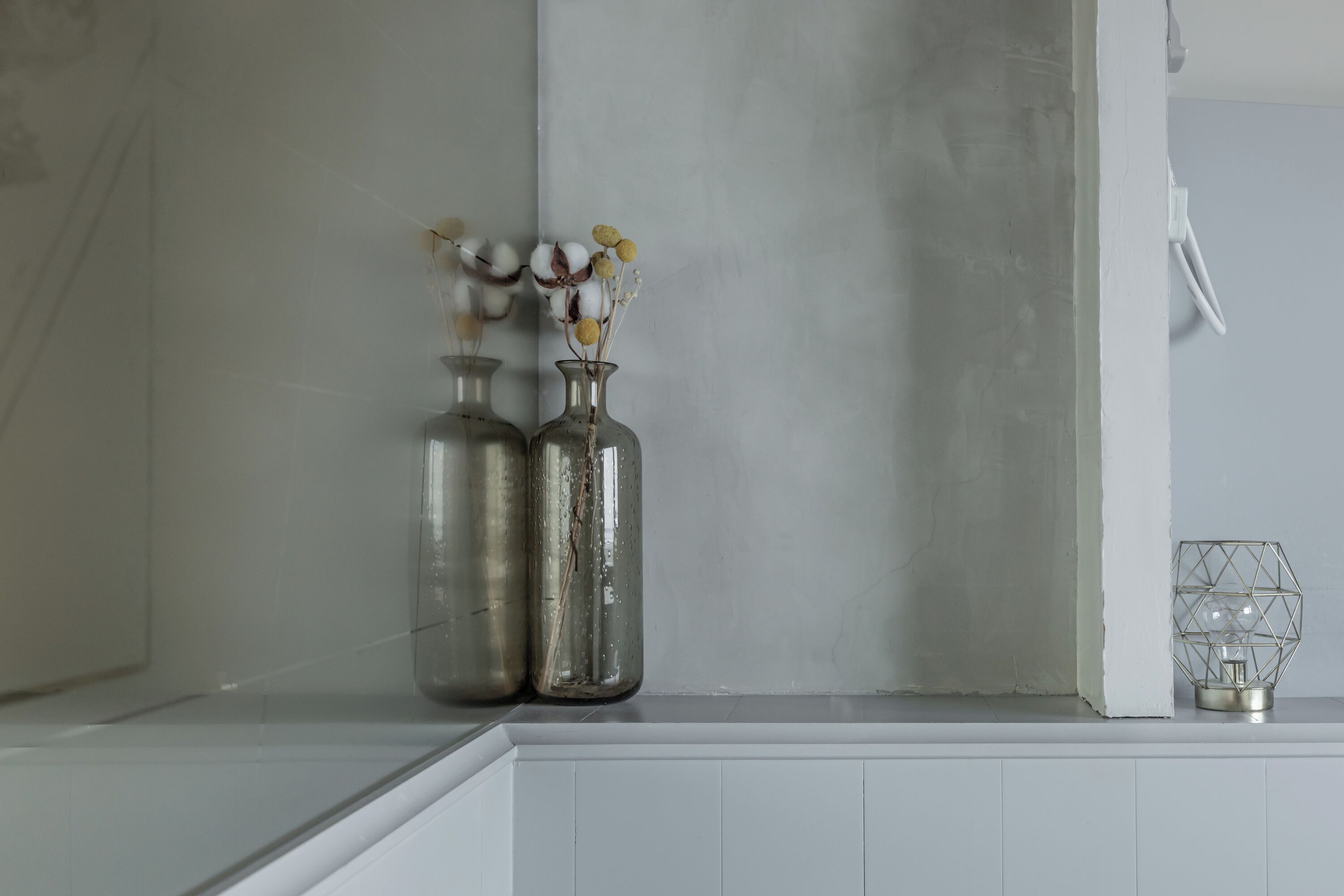 Two metal vases with dried flowers on a white shelf against a plain wall.
