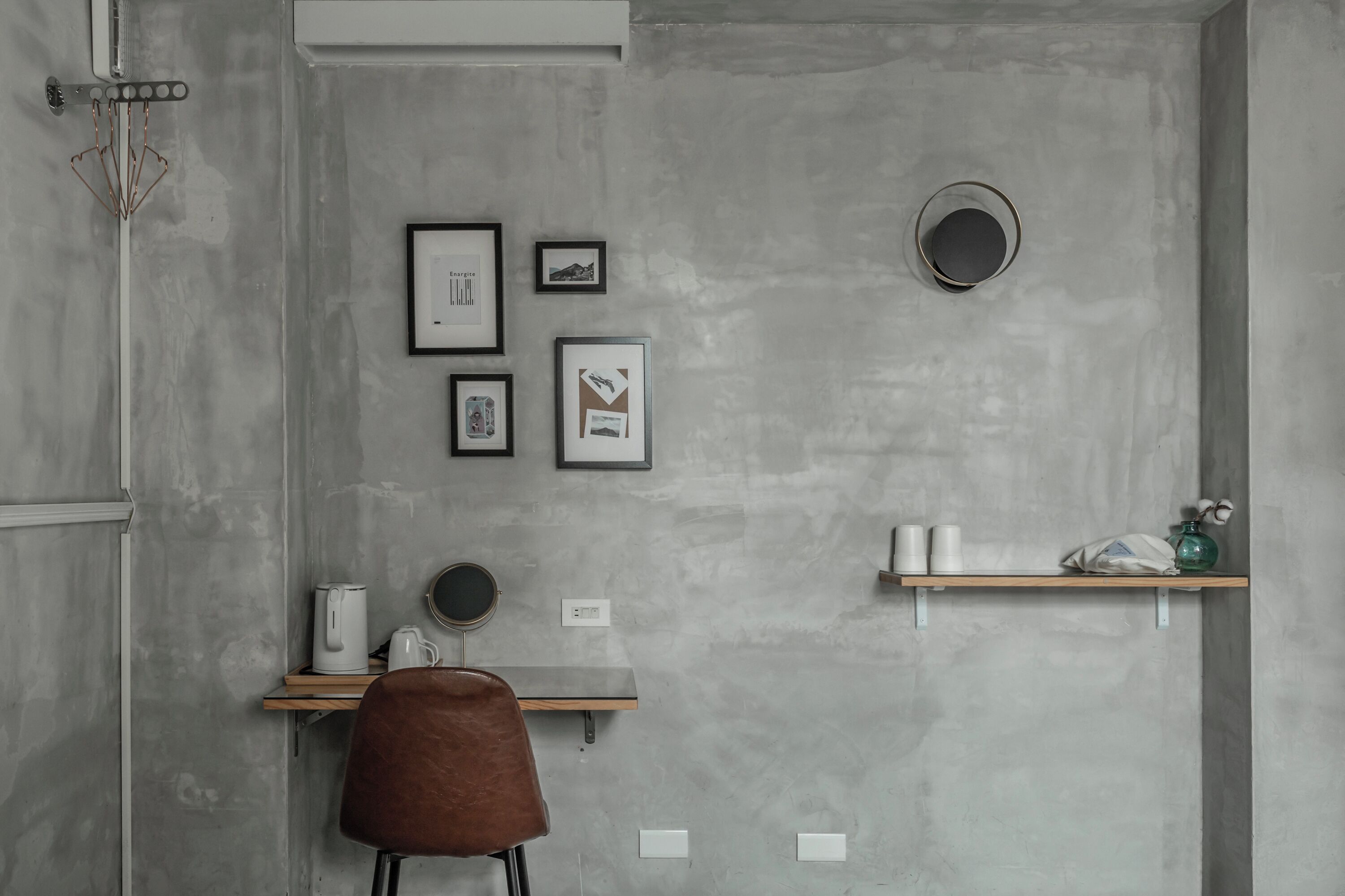 A modern, minimalist room with a concrete wall, a wooden desk, a brown chair, and a shelf with toiletries.