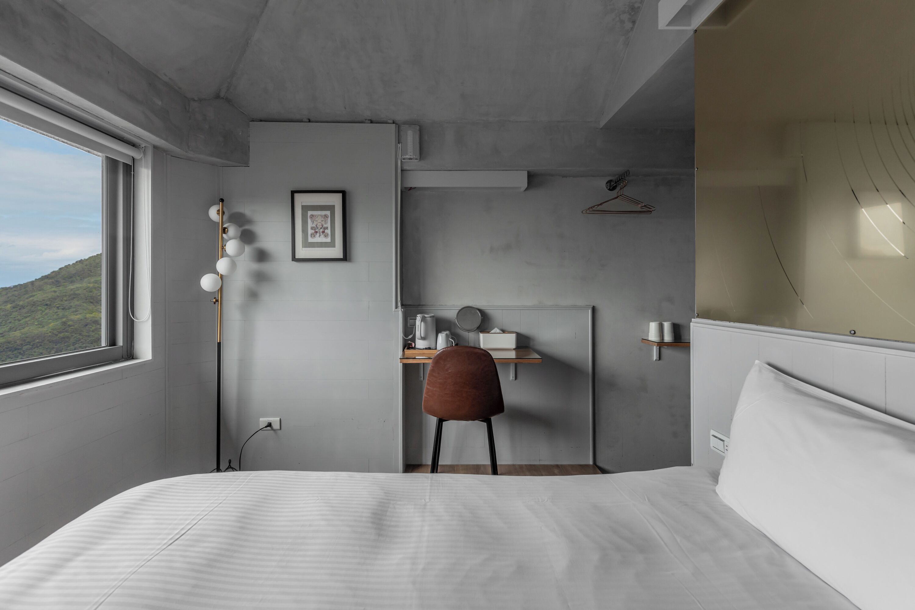 A modern bedroom with a large bed, a desk with a chair, a hanging light fixture, and a view of the outdoors.