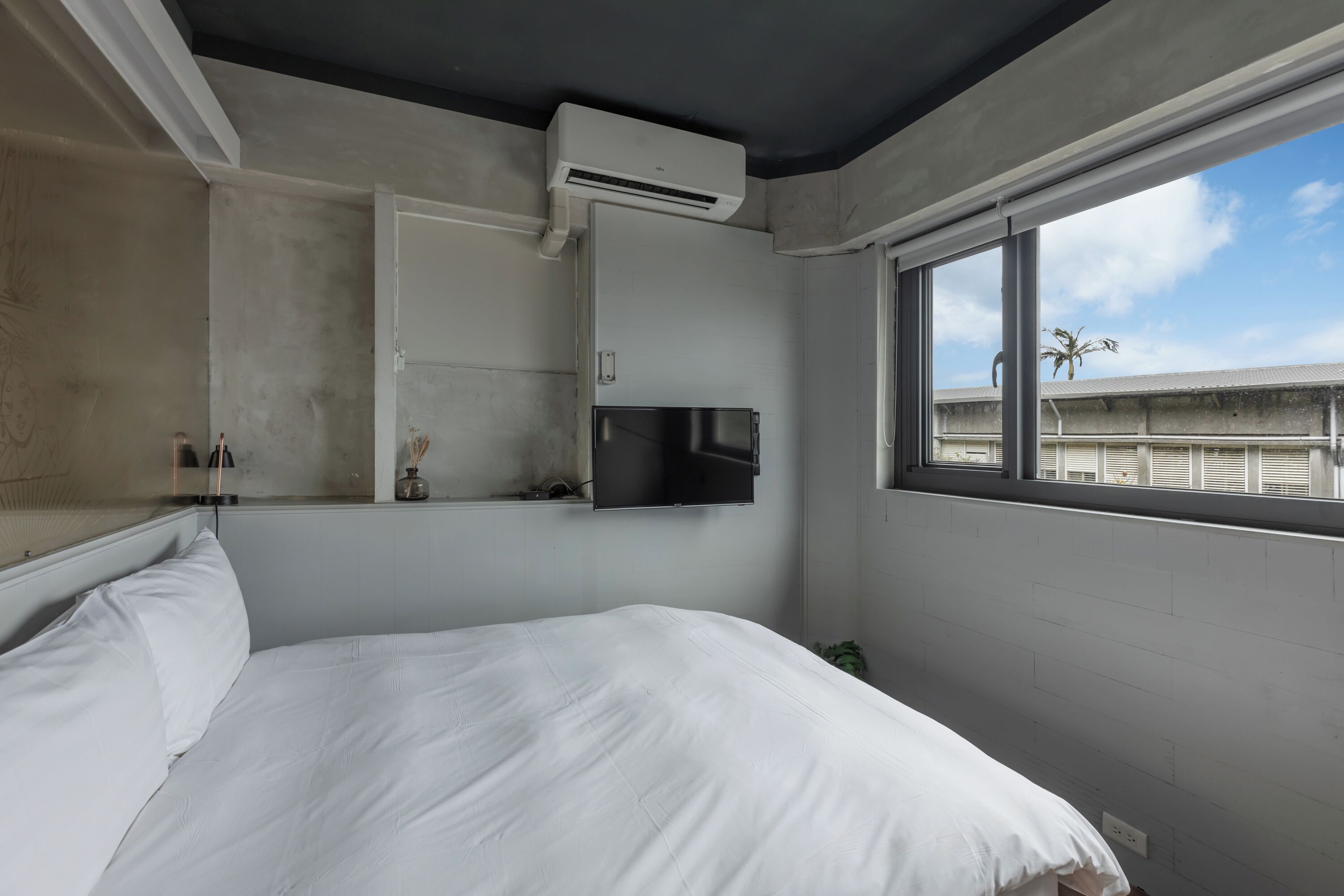 Une chambre d’hôtel équipée d’un lit, d’un climatiseur, d’un téléviseur à écran plat fixé au mur et d’une fenêtre donnant sur un immeuble et un palmier.