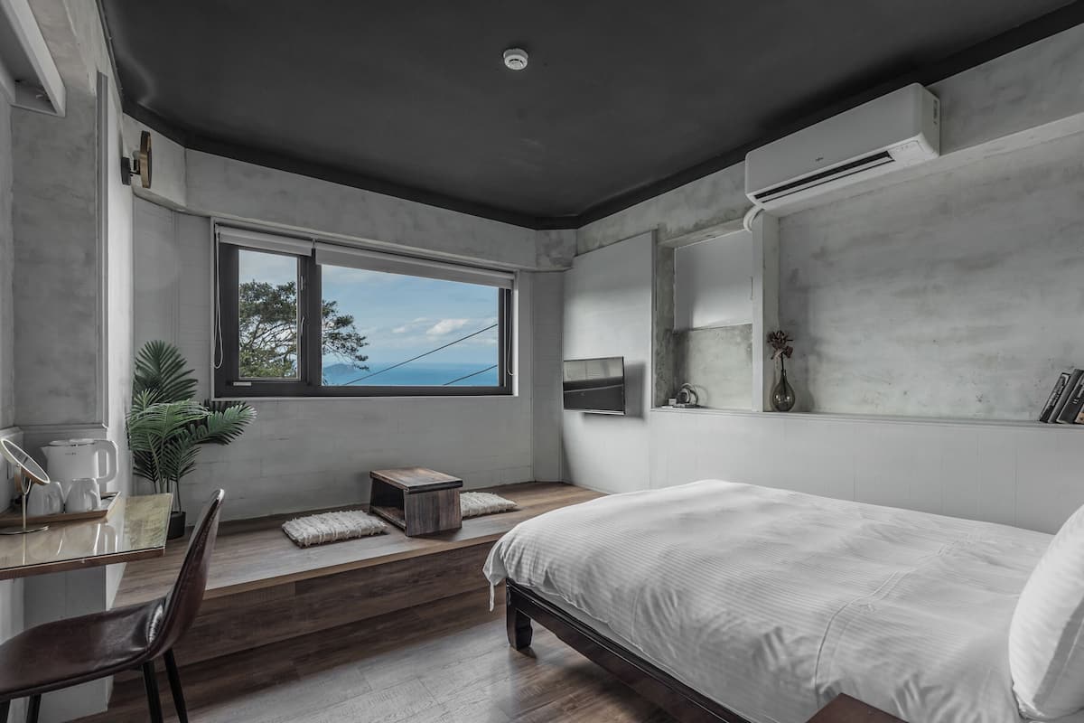 Un dormitorio moderno con una cama grande, un escritorio con silla, una ventana con vistas y una pequeña mesa con una planta.