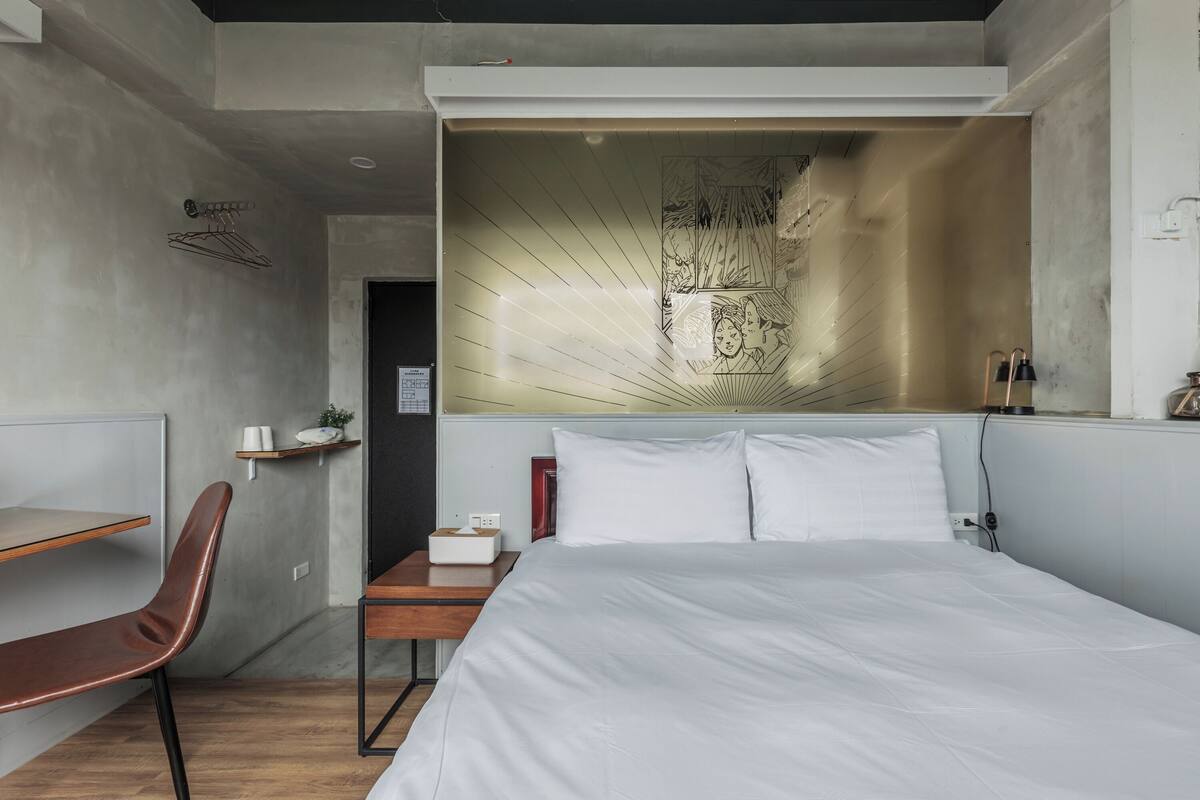 Una habitación de hotel moderna con una cama grande, un escritorio y una silla. Se observa una estantería en la pared con una planta y una caja de pañuelos.