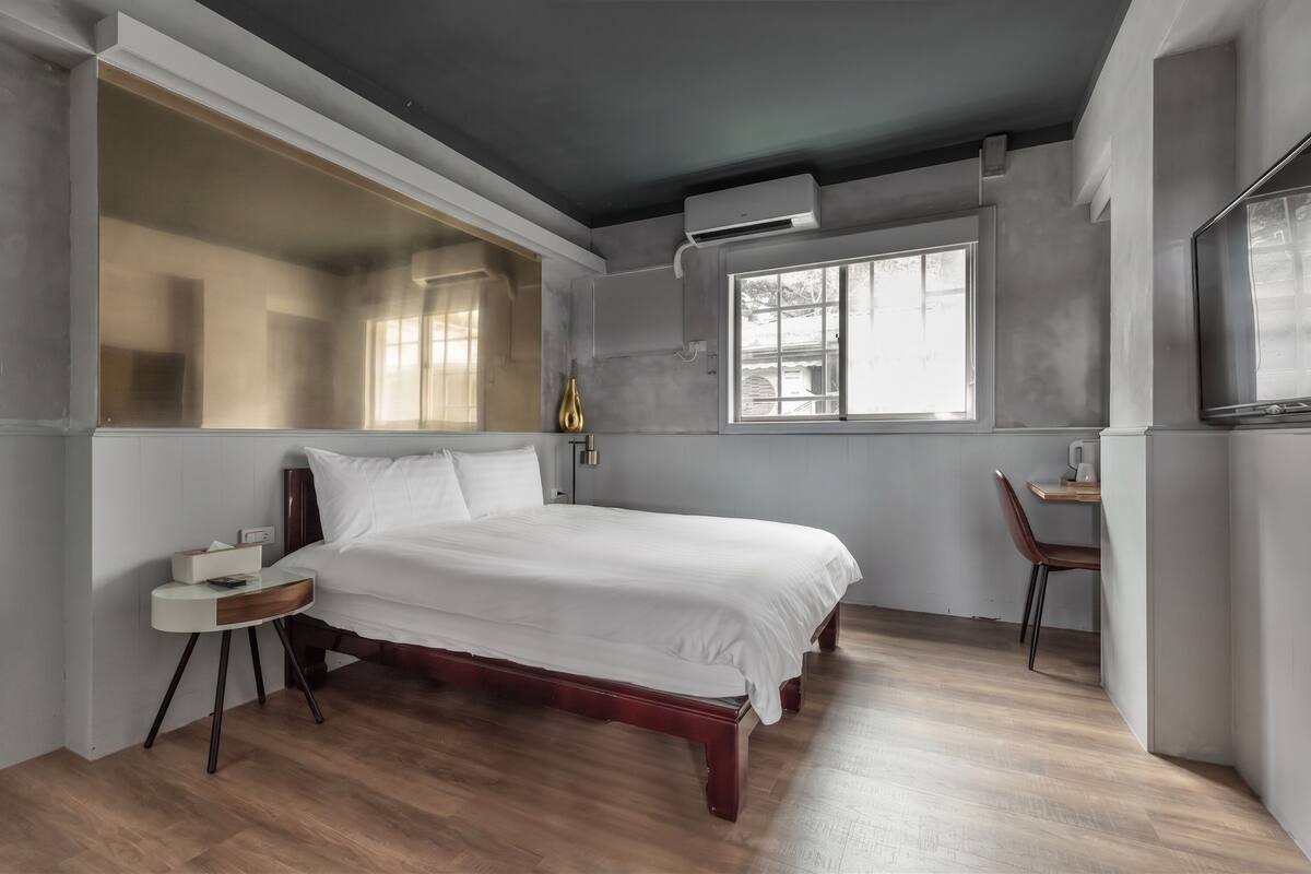 Un dormitorio con cama, mesita de noche, silla, ventana y aire acondicionado.