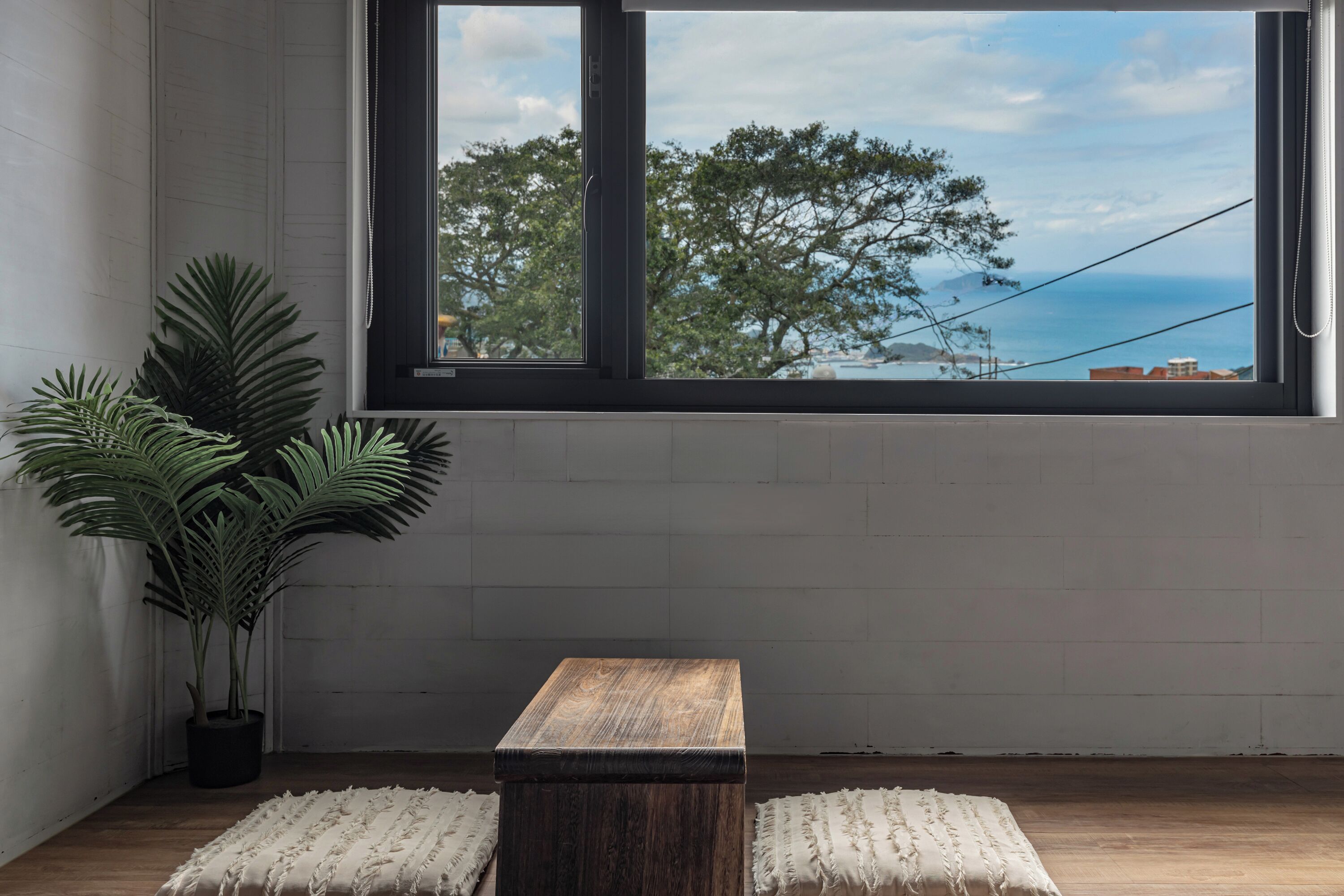 A room with a large window, a wooden table, two white poufs, and a potted plant.