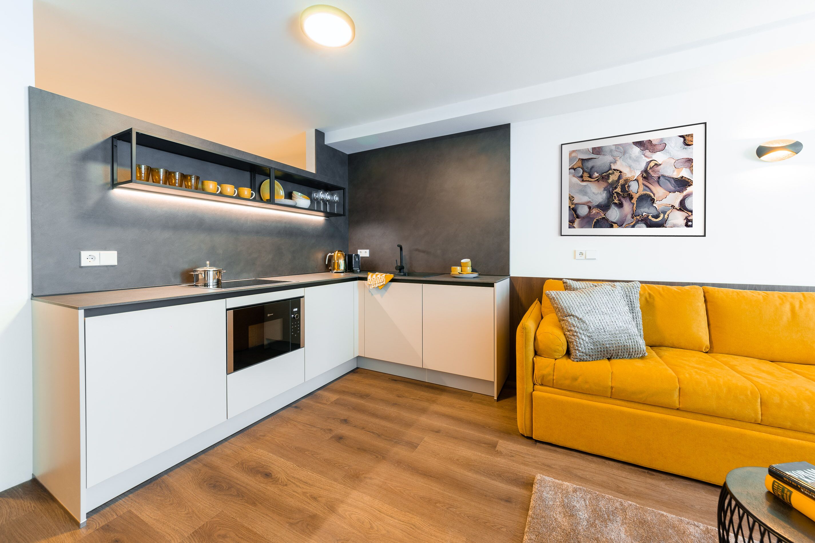 A modern kitchen with white cabinets, a built-in oven, and a coffee machine. There's a yellow sofa with a throw pillow and a framed abstract painting on the wall.