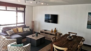 A modern living room with a gray sofa, patterned armchairs, a wooden coffee table, a dining table with chairs, a flat-screen TV mounted on the wall, and a large window with blinds. - Witsand 806 (Cape Town)