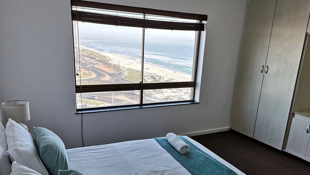 A hotel room with a large window offering a view of a beach and road. The room has a bed with white linens, a teal throw blanket, and a rolled towel. - Witsand 806 (Cape Town)