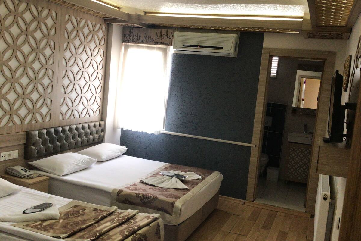A hotel room with two beds, a decorative headboard, an air conditioning unit, and a view of the cityscape.