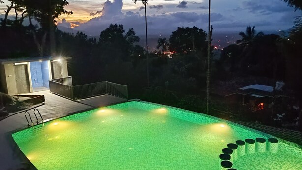 Una piscina con un deck iluminado en verde, una estructura pequeña adyacente con un muro de azulejos azules, y vista a una ciudad al atardecer.