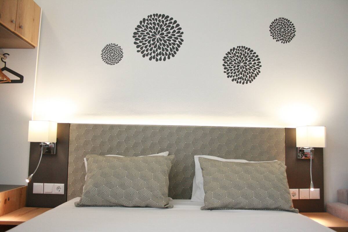Una habitación de hotel con una cama bien hecha, dos almohadas estampadas, un cabecero con diseño geométrico y lámparas fijadas a la pared.