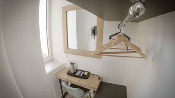Ein kleines Zimmer mit Schreibtisch aus Holz, einem Stuhl, einem hängenden Holzanzhänger, einem Spiegel und einer Deckenleuchte.