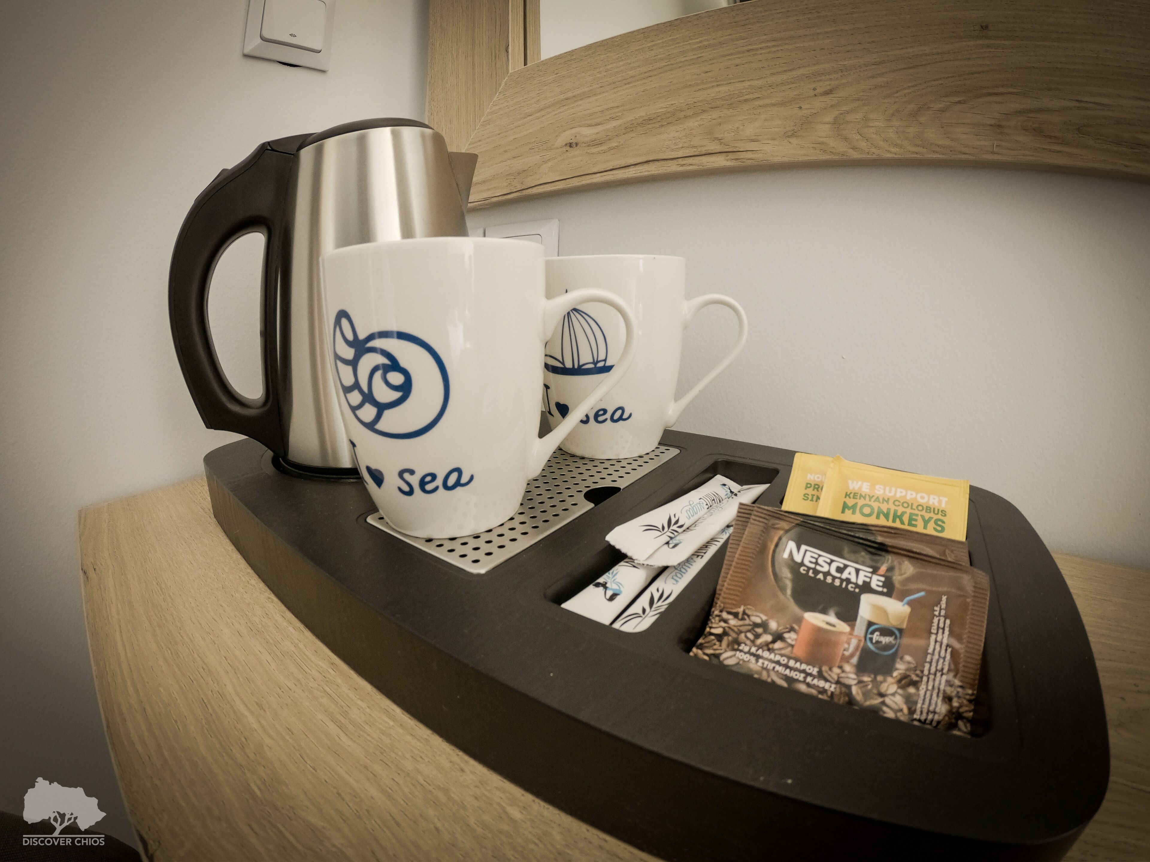 A coffee station with a coffee maker, two cups with 'sea' branding, and a tray with coffee pods and a packet of Nescafe Classic.