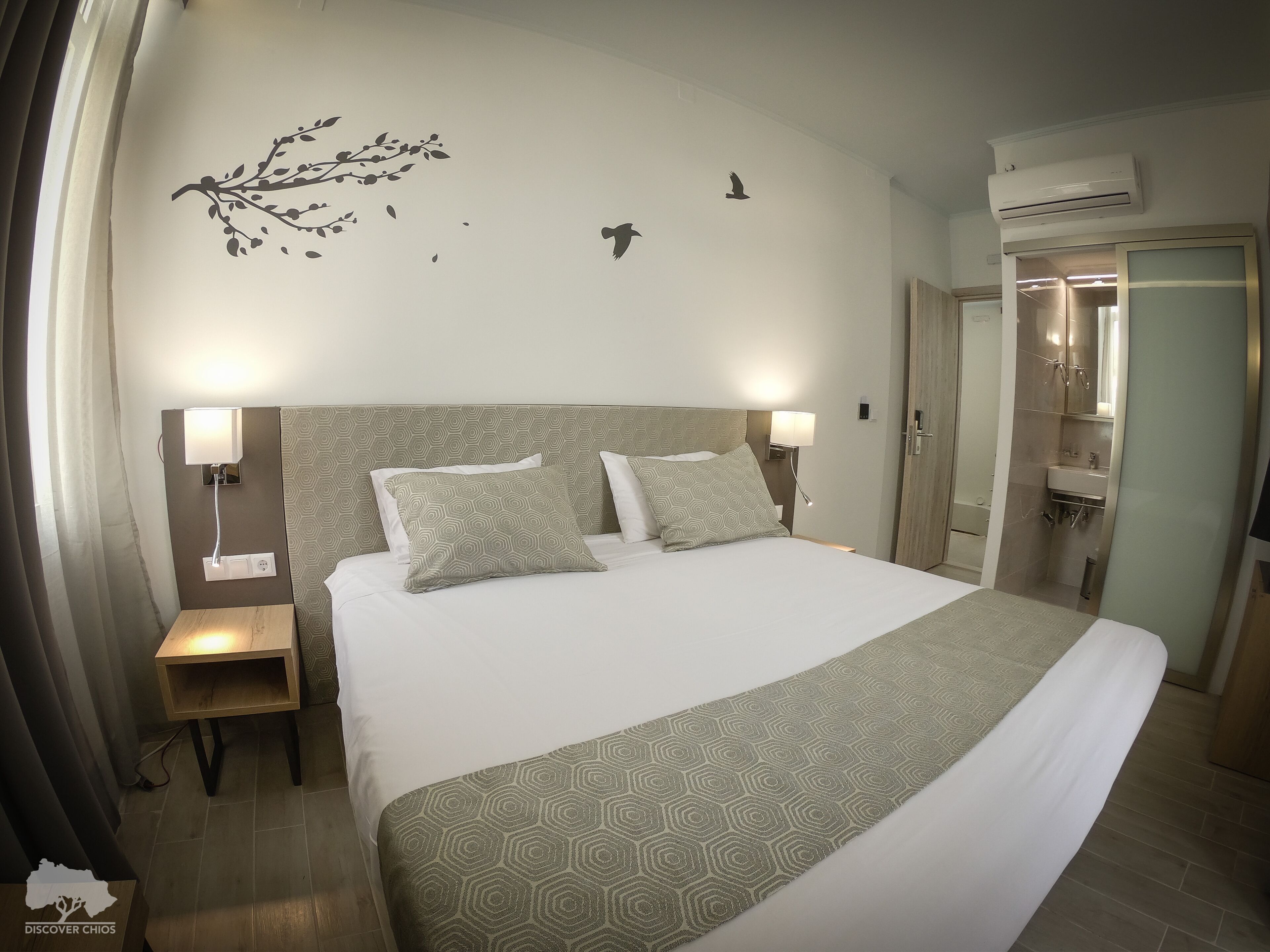 Ein Hotelzimmer mit einem großen Bett, zwei Nachttischen, einer Wand-Klimaanlage und einem durch eine offene Tür einsehbaren Badezimmer.