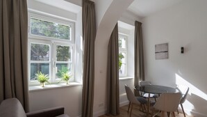 Ein modernes Wohnzimmer mit Glastisch und Stühlen, großen Fenstern mit Vorhängen und Blick ins Grüne.