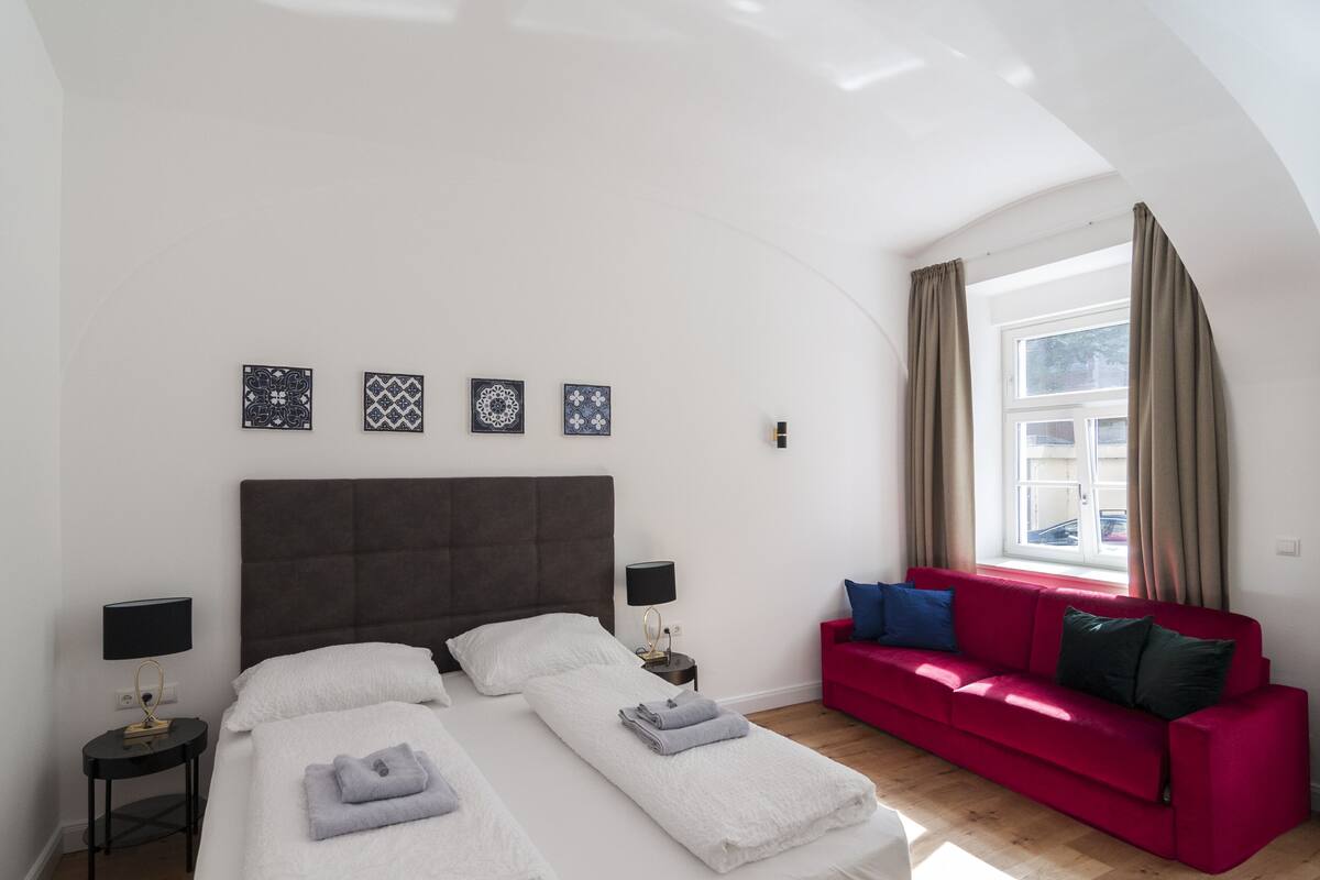 Quarto com duas camas, um sofá vermelho e uma janela com cortinas.