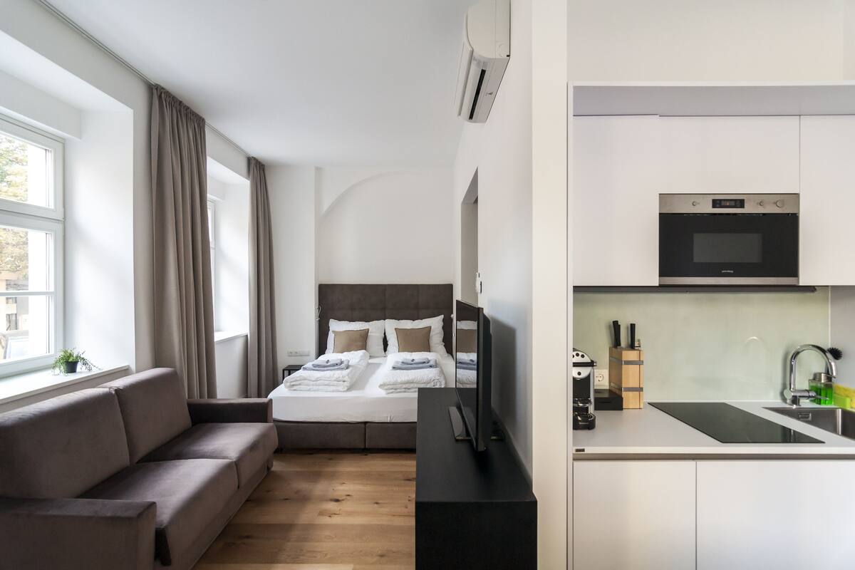 Quarto de hotel moderno com sofá, cama com roupa branca, televisão de tela plana, área de cozinha com micro-ondas e pia.