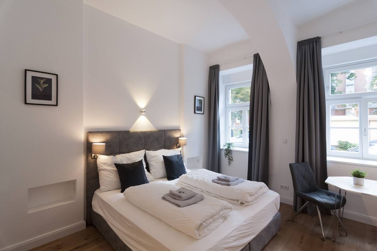 Quarto de hotel moderno com uma cama grande, abajures, uma mesinha com uma planta e vista para o exterior pela janela.