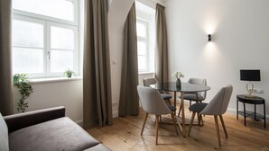 Ein modernes Wohnzimmer mit einem runden Esstisch, grauen Stühlen, einer Couch, einem Fernseher auf einem Schrank und einem Fenster mit Vorhängen.