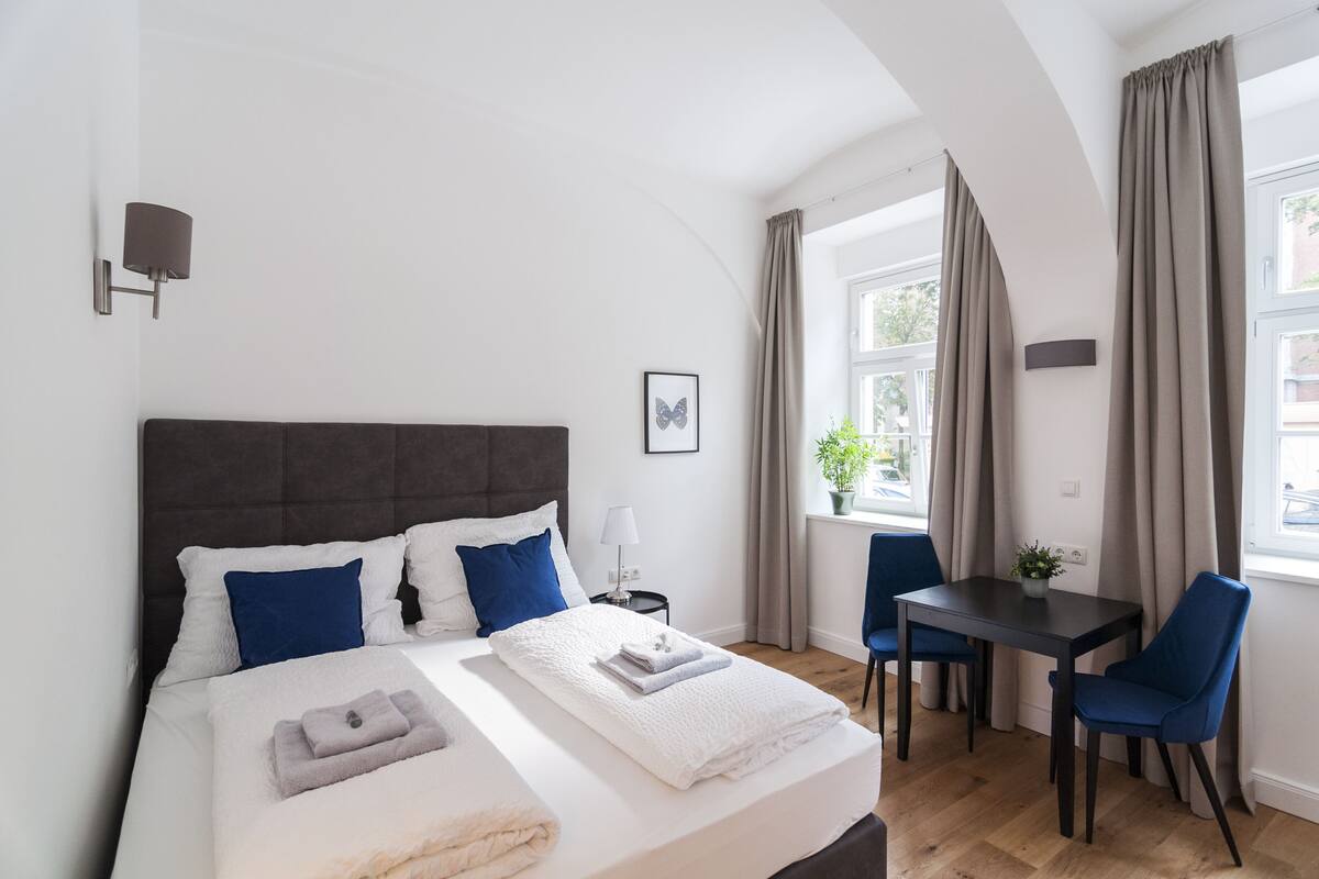 Quarto moderno com cama grande, uma mesa pequena, duas cadeiras e janela com cortinas.