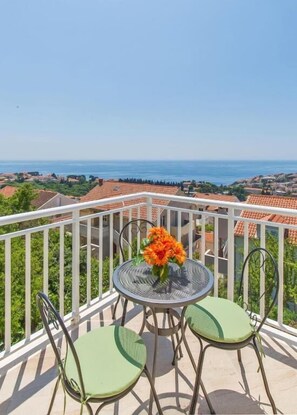 A balcony with a round table and two chairs, overlooking a coastal town with terracotta roofs and a clear blue sky. - Rooms on Konalu (Dubrovnik)