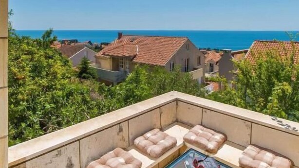 A balcony with cushioned seating, a glass table with a cake, and a view of the sea and buildings. - Rooms on Konalu (Dubrovnik)