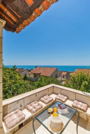 A balcony with cushioned seating, a glass table with a cake, and a view of the sea and buildings. - Rooms on Konalu (Dubrovnik)