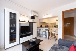 A modern living room with a flat-screen TV, a coffee table, and a sofa. There is a kitchen area with bar stools and a microwave.